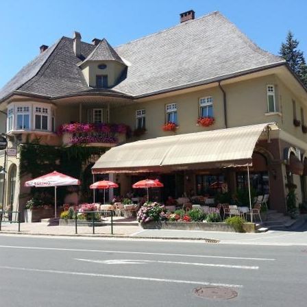 Ein großes gelbes Gebäude mit einem grauen Dach und einem grünen Vordach. Das Gebäude hat mehrere Fenster und Blumenkästen. Draußen stehen Tische und Stühle unter Schirmen.