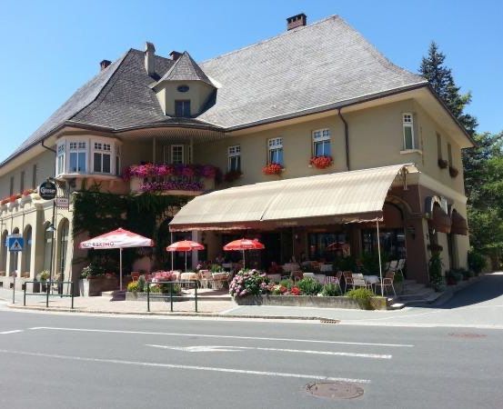 Ein großes gelbes Gebäude mit einem grauen Dach und einem grünen Vordach. Das Gebäude hat mehrere Fenster und Blumenkästen. Draußen stehen Tische und Stühle unter Schirmen.