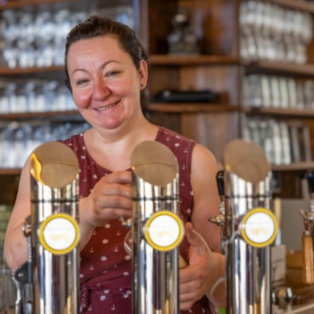 Eine Frau steht hinter der Theke in einer Bar, lächelt und hält einen Bierhahn.