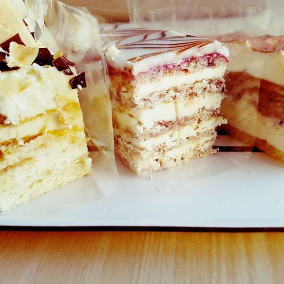 Nahaufnahme mehrerer geschichteter Kuchen auf einem weißen Teller, teilweise mit Plastikfolie bedeckt. Die Kuchen haben verschiedene Beläge wie Schokolade, Sahne und Beeren.