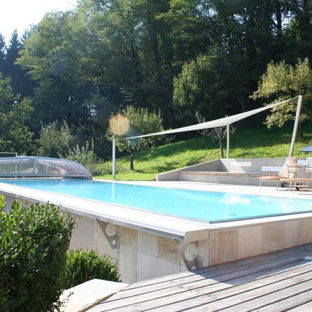 Ein leerer Swimmingpool befindet sich auf einer hölzernen Terrasse mit üppiger Grünfläche im Hintergrund. Darüber befindet sich ein weißes Vordach.