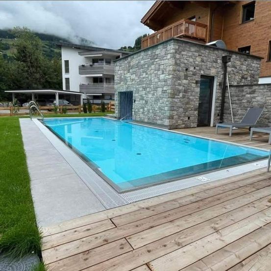 Ein Freischwimmbecken mit Holzdeck ist von einer Steinmauer und einem Gebäude mit Balkonen umgeben. Zwei Liegestühle stehen auf dem Deck.