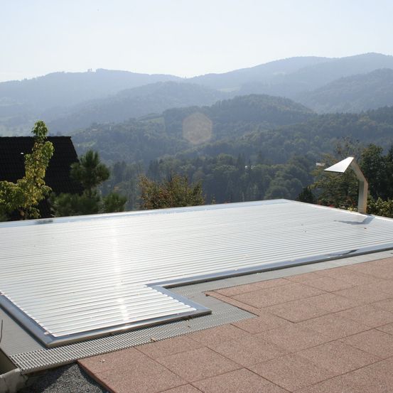 Dach mit Metallbedeckung und einem Solarpanel. Im Hintergrund befinden sich Berge und Bäume. Der Himmel ist klar und blau.