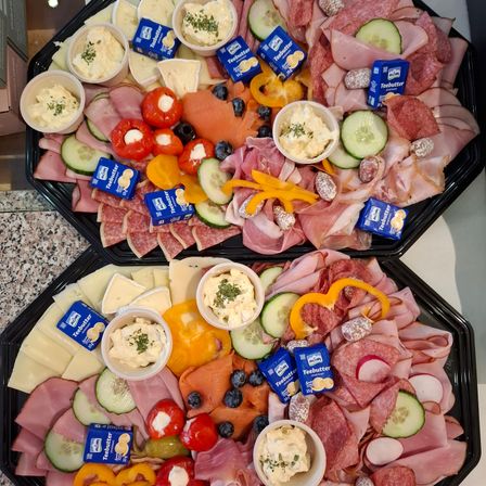 Zwei schwarze Tabletts enthalten eine Auswahl an Lebensmitteln: geschnittenes Fleisch, Käse, Gemüse und Dips. Jedes Tablett hat geschnittene Gurken, Paprika, Oliven und kleine Tassen mit Dips. Es gibt blaue und gelbe Packungen 'Teebutter' Aufstrich.