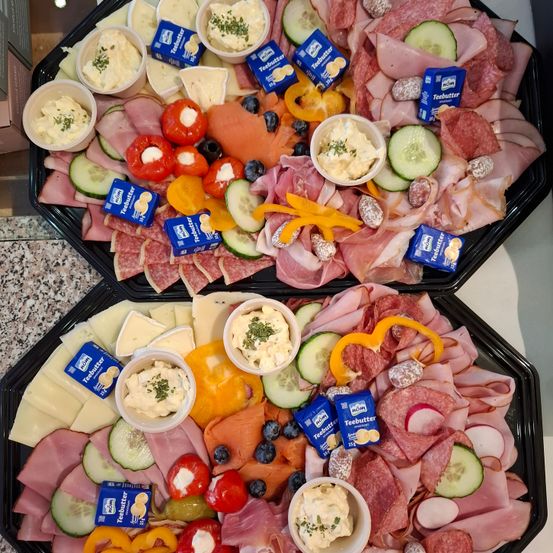 Zwei schwarze Tabletts enthalten eine Auswahl an Lebensmitteln: geschnittenes Fleisch, Käse, Gemüse und Dips. Jedes Tablett hat geschnittene Gurken, Paprika, Oliven und kleine Tassen mit Dips. Es gibt blaue und gelbe Packungen 'Teebutter' Aufstrich.