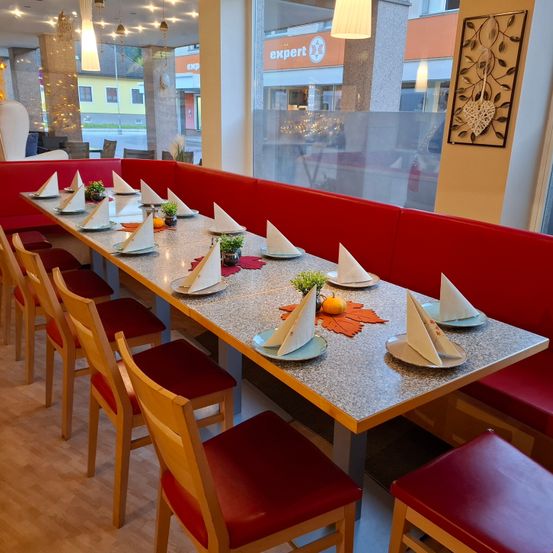 Ein Restaurant mit einem langen Tisch für Gäste. Der Tisch hat Teller, Servietten und dekorative Blätter. Die Stühle sind rot mit Ledersitzen. Die Wände sind weiß und es gibt ein großes Fenster.