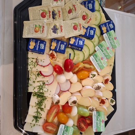 Ein schwarzer Teller zeigt eine Auswahl an Käse, Gemüse und Beilagen. Verschiedene Käsescheiben werden von Tomaten, Radieschen und Gurken begleitet. Senf und Butter gehören ebenfalls dazu.