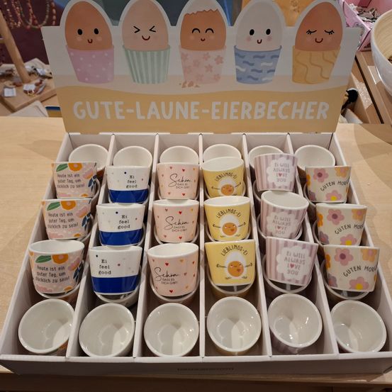 Eine Schachtel mit vielen kleinen Bechern mit fröhlichen Cartoon-Eiern darauf, der Text lautet 'Gute-Laune-Eierbecher'. Einige Becher sagen 'Ich will immer dich lieben', andere sagen 'Ich fühle mich gut'. Leere Becher sind ebenfalls enthalten.