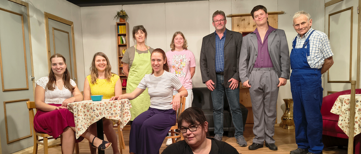 Eine Gruppe von Menschen in verschiedenen Kostümen posiert für ein Foto in einem Raum mit Holzböden und weißen Wänden. Es gibt einen Tisch mit einer blumigen Tischdecke und einer Schüssel darauf.