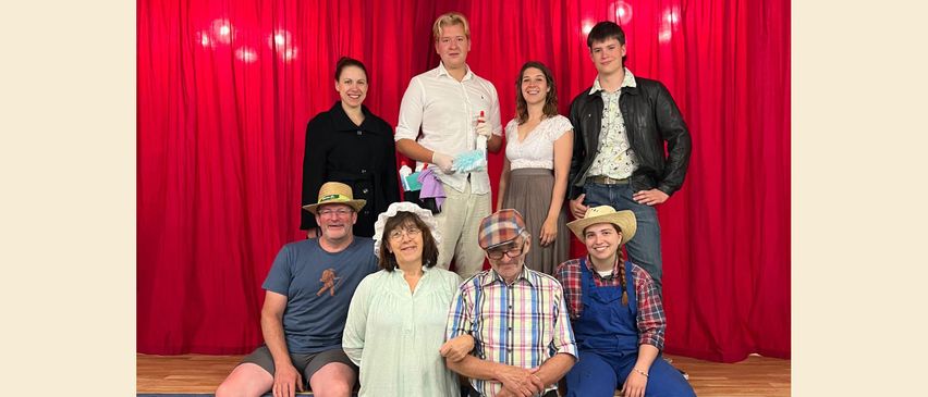 Eine Gruppe von Menschen in verschiedenen Kostümen posiert vor einem roten Vorhang. Sie scheinen Schauspieler oder Teilnehmer einer Aufführung oder Veranstaltung zu sein.