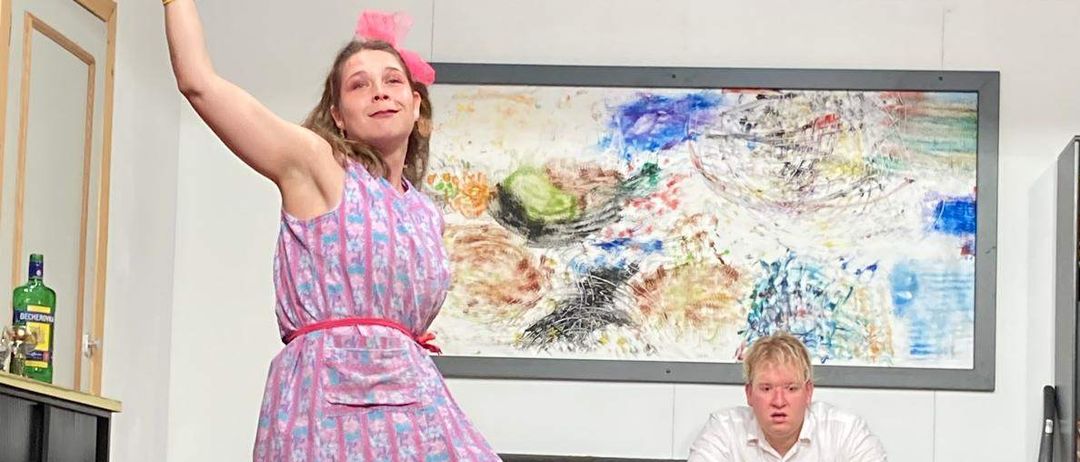 Eine Frau in einem rosa Kleid und Handschuhen steht auf der Bühne und hält einen Kolben. Ein Mann sitzt auf einem Sofa im Hintergrund. Ein abstraktes Gemälde hängt an der Wand.