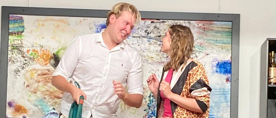 Ein Mann mit einem Staubsauger steht neben einer Frau in einem Leopardenmuster-Outfit, beide lächeln und sehen sich in einem Raum an.
