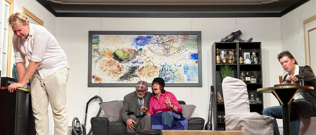 Eine Bühnenaufstellung mit einem Paar auf einer Couch, einer Person in der Nähe eines Staubsaugers, einem Regal mit Flaschen und einem gerahmten Gemälde an der Wand.