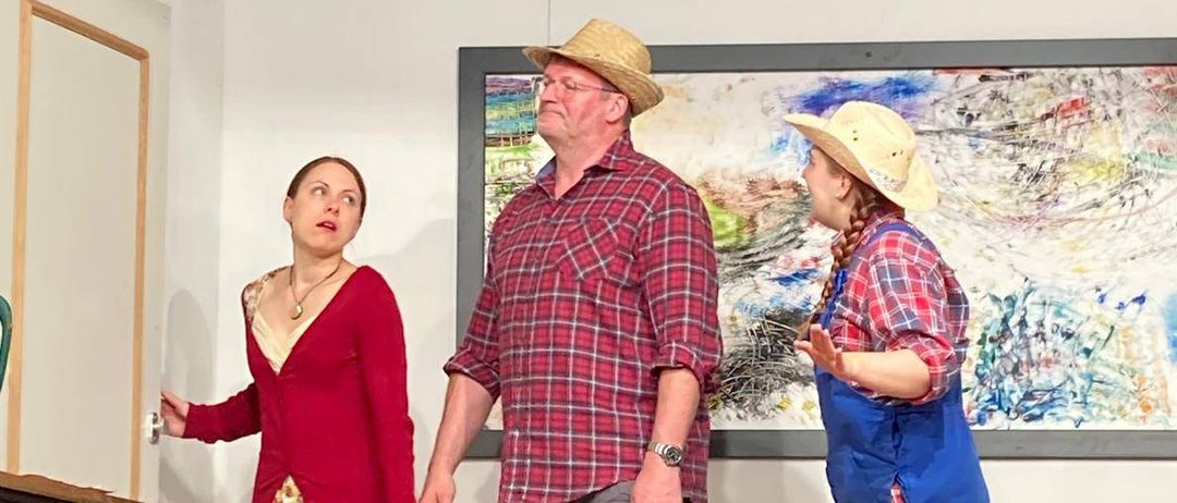 Drei Personen stehen in einem Innenraum auf einem Holzboden. Eine Frau in einem Blumenrock und einem roten Pullover öffnet eine Tür. Ein Mann in einem karierten Hemd und einem Hut steht hinter ihr. Eine weitere Frau in einer blauen Arbeitshose und Stiefeln steht rechts. Hinter ihnen befindet sich eine Couch mit Kissen, ein gerahmtes Gemälde und ein Mülleimer.