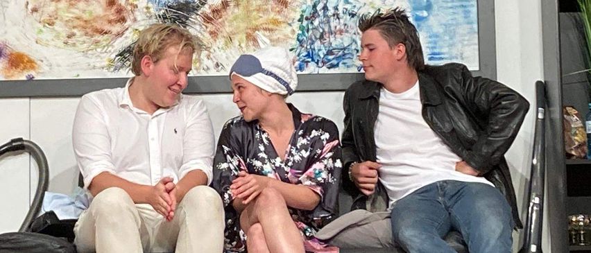 Drei Personen sitzen auf einer Couch, zwei Männer lächeln und eine Frau trägt eine Kappe. Hinter ihnen befindet sich ein abstraktes Gemälde.