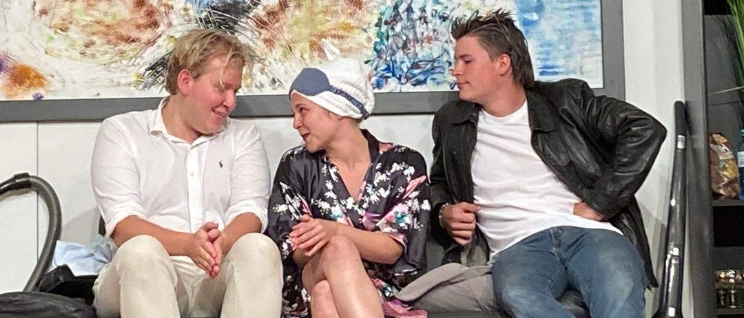 Drei Personen sitzen auf einer Couch, zwei Männer lächeln und eine Frau trägt eine Kappe. Hinter ihnen befindet sich ein abstraktes Gemälde.
