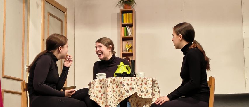 Drei Frauen sitzen um einen Tisch herum und unterhalten sich. Der Tisch hat eine Tischdecke mit Blumenmuster und einen Teekessel. Sie befinden sich in einem Raum mit Holzboden und weißen Wänden.