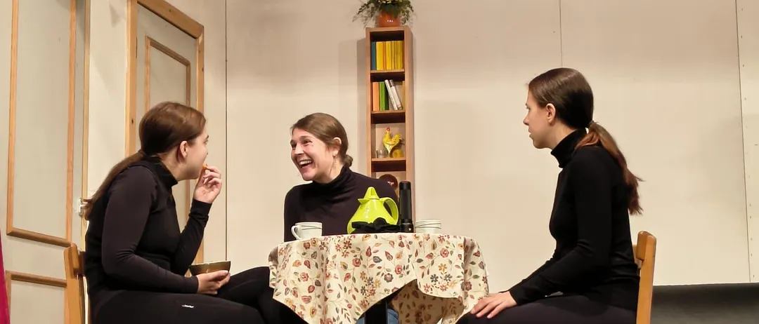 Drei Frauen sitzen um einen Tisch herum und unterhalten sich. Der Tisch hat eine Tischdecke mit Blumenmuster und einen Teekessel. Sie befinden sich in einem Raum mit Holzboden und weißen Wänden.