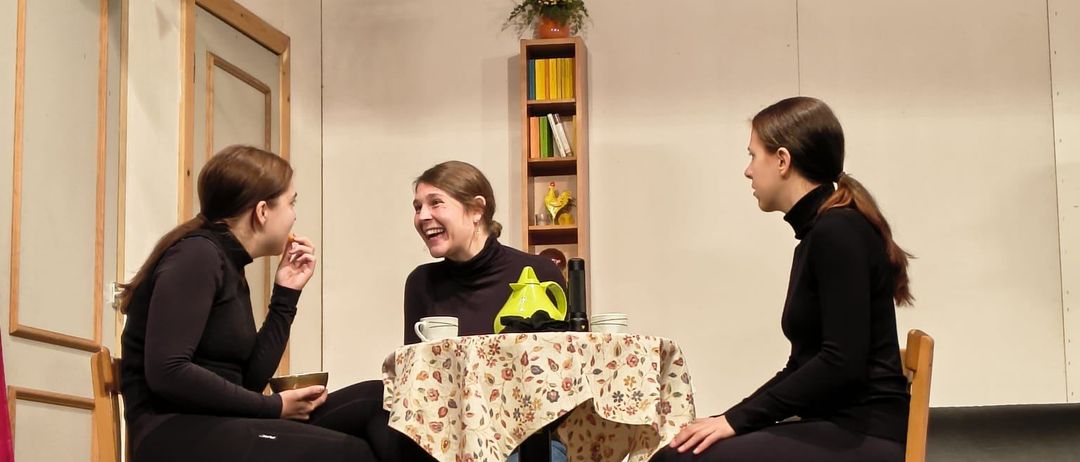 Drei Frauen sitzen um einen Tisch herum und unterhalten sich. Der Tisch hat eine Tischdecke mit Blumenmuster und einen Teekessel. Sie befinden sich in einem Raum mit Holzboden und weißen Wänden.