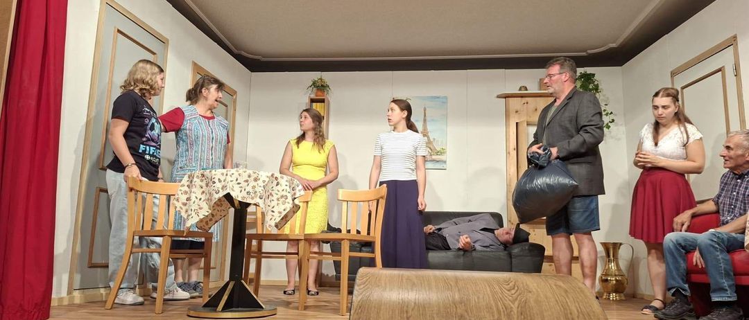 Eine Gruppe von Menschen steht auf der Bühne, möglicherweise bei einer Aufführung. Eine Frau in einem gelben Kleid und ein Mann in einem Anzug stehen neben einem Tisch. Ein anderer Mann liegt auf einem Sofa.