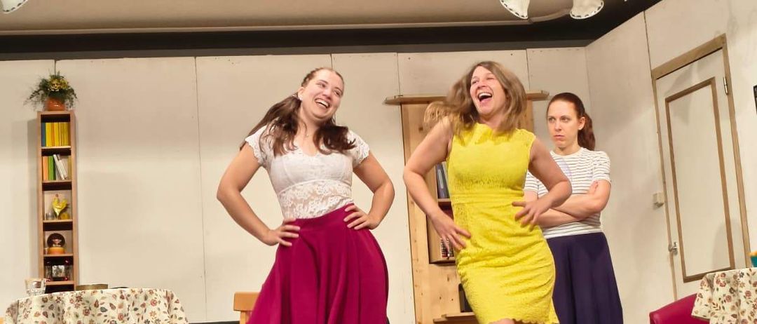 Drei Frauen auf der Bühne, eine in einem gelben Kleid und eine andere in einem rosa Rock, lachen zusammen. Dahinter steht ein Tisch mit einer Tischdecke mit Blumenmuster, ein Bücherregal und eine Couch. Ein Lautsprecher und eine Tür sind ebenfalls sichtbar.