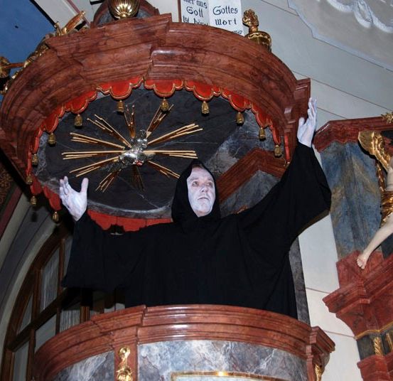 Eine Person in einem schwarzen Gewand steht in einer Kirche mit erhobenen Armen in der Nähe einer goldenen Statue.