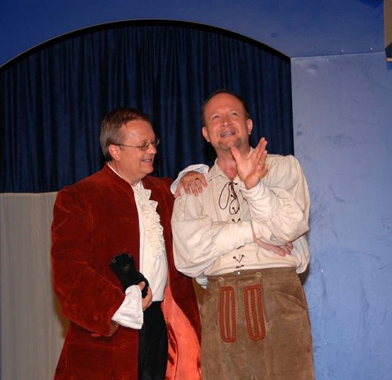 Zwei Männer stehen auf der Bühne, einer in einem roten Mantel und der andere in einem weißen Hemd mit brauner Hose. Sie scheinen zu applaudieren.