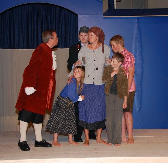 Eine Gruppe von Menschen in Kostümen auf einer Bühne, wahrscheinlich bei einer Aufführung. Ein Mann in einem roten Mantel steht links.