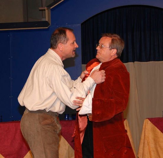 Zwei Männer sind in einer theatralischen Aufführung auf der Bühne engagiert, einer passt die Kleidung des anderen an. Die Umgebung umfasst Tische mit roten und gelben Tüchern und blauen Schalen.