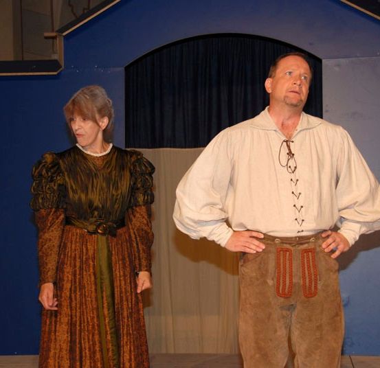 Ein erwachsener Mann und eine Frau stehen auf der Bühne und schauen sich an. Die Frau trägt ein langes, gemustertes Kleid. Der Mann trägt ein weißes Hemd mit Schnürsenkeln und braune Hosen.