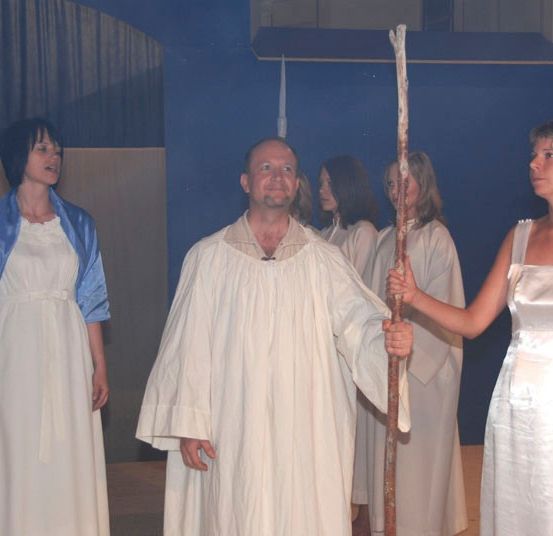 Ein Mann in einer weißen Robe hält einen Stab und steht zwischen zwei Frauen in weißen Kleidern. Sie sind auf einer Bühne mit blauem Hintergrund.