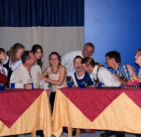 Eine Gruppe von Erwachsenen und Kindern versammelt sich um zwei Tische mit blauen Tassen und Tellern in einem Raum mit blauem Hintergrund und Vorhängen.