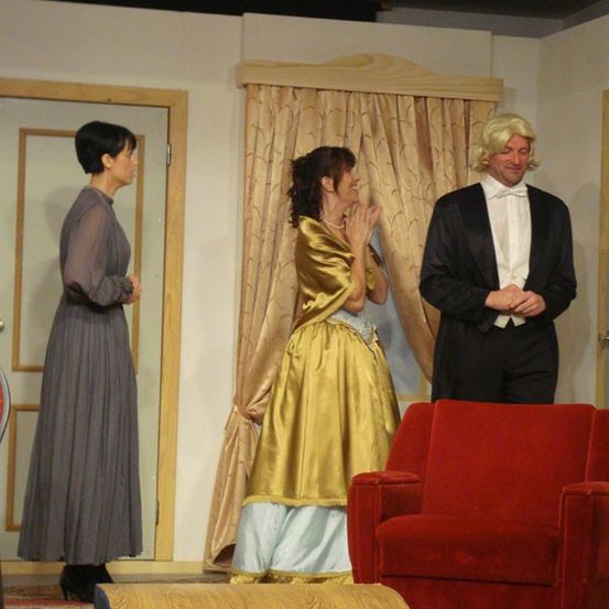 Drei Menschen in Kostümen stehen auf einer Bühne. Eine Frau in einem gelben Kleid steht einem Mann in einem schwarzen Frack gegenüber. Eine weitere Frau in einem grauen Kleid steht in der Nähe einer Tür.
