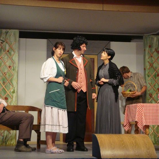 Fünf Personen stehen auf einer Bühne und führen ein Gespräch. Ein Mann sitzt auf einer Bank in der Nähe. Ein Tisch mit einem Korb und einem Blumenvase steht rechts.