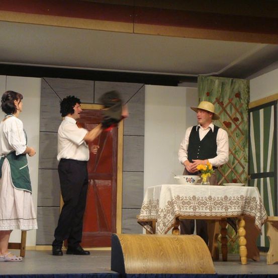 Drei Personen auf der Bühne, ein Mann hält einen Handschuh, ein anderer in einem Hut schaut ihn an und eine Frau in einer grünen Schürze. Hinter ihnen steht ein Tisch mit einer Schüssel und Blumen und eine Holztür.