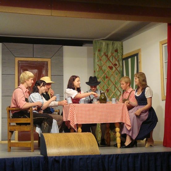 Eine Gruppe von Menschen sitzt auf der Bühne um einen Tisch mit Tassen und einer Kanne. Sie scheinen in einem Theaterstück zu spielen.