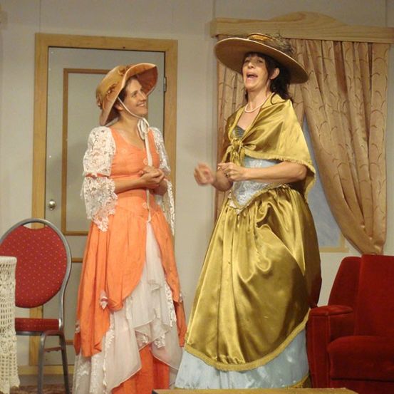 Zwei Frauen in aufwendigen Kostümen aus der Vergangenheit stehen auf der Bühne, wahrscheinlich bei einer Probe für ein Theaterstück. Eine trägt ein oranges Kleid, die andere ein goldenes Gewand. Im Hintergrund befinden sich eine Tür und rote Stühle.