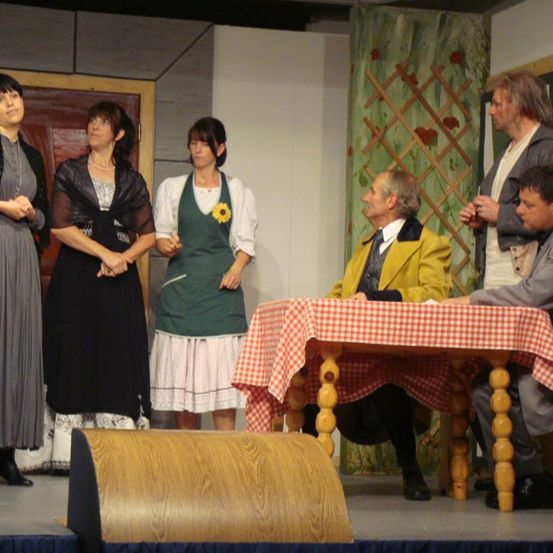 Fünf Personen stehen auf der Bühne, möglicherweise in einem Theaterstück. Zwei Frauen und ein Mann stehen, während zwei Männer an einem Tisch sitzen. Der Tisch hat eine rot-weiß karierte Tischdecke.