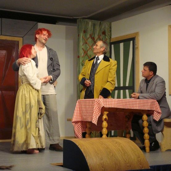 Drei Personen stehen auf der Bühne, zwei stehend und eine sitzend. Der Mann in der Mitte trägt einen gelben Mantel und hält einen Stift. Die Frau im gelben Kleid schaut ihn an. Der andere Mann beobachtet sie. Der Raum hat Holztüren und eine Holzbank.