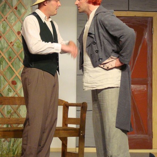 Zwei Männer stehen auf der Bühne. Einer trägt einen Strohhut und eine Weste, der andere hat rote Haare und eine graue Strickjacke. Sie scheinen sich zu unterhalten. Hinter ihnen steht eine hölzerne Bank.