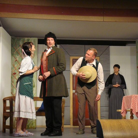 Auf der Bühne stehen mehrere Schauspieler in Kostümen. Ein Mann hält einen Strohhut. Eine Frau in einer Schürze steht neben einem Mann in Uniform. Eine andere Frau steht bei einem Tisch mit rot-karierter Tischdecke.