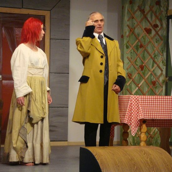 Ein Mann in einem gelben Mantel steht auf der Bühne mit einer Frau, beide schauen in eine Richtung. Hinter ihnen steht eine rote Tür und ein Tisch mit einer rot-weiß karierten Tischdecke.