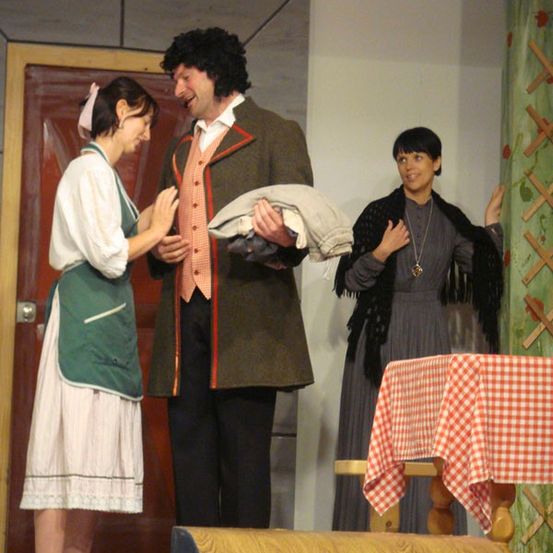 Drei Personen stehen auf der Bühne, eine hält ein gefaltetes Kleidungsstück. Die Frau in Grün lächelt und spricht mit dem Mann im grünen Mantel. Dahinter steht eine Frau mit einer Halskette neben einem Tisch mit einer rot-weiß karierten Tischdecke.