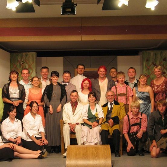 Eine Gruppe von Menschen in verschiedenen Kostümen, einige sitzend und andere stehend, posiert für ein Foto in einem Theater.