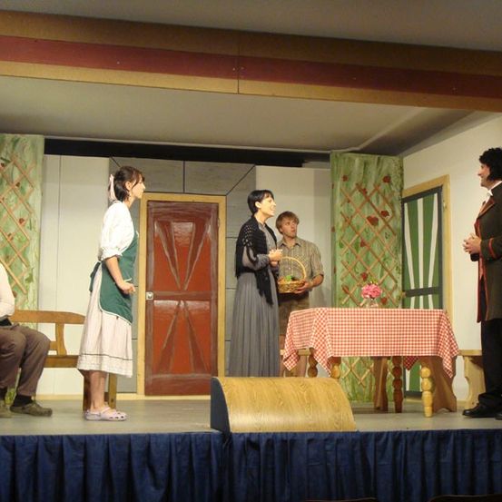 Fünf Personen stehen auf einer Bühne mit einer Holztür, einem Tisch und einer Bank. Ein Mann mit Hut sitzt auf der Bank. Eine Frau hält einen Korb. Eine weitere Frau hält eine Blumenvase. Ein Mann steht neben einer Frau mit einem Korb. Ein Mann steht neben einem roten Vorhang.