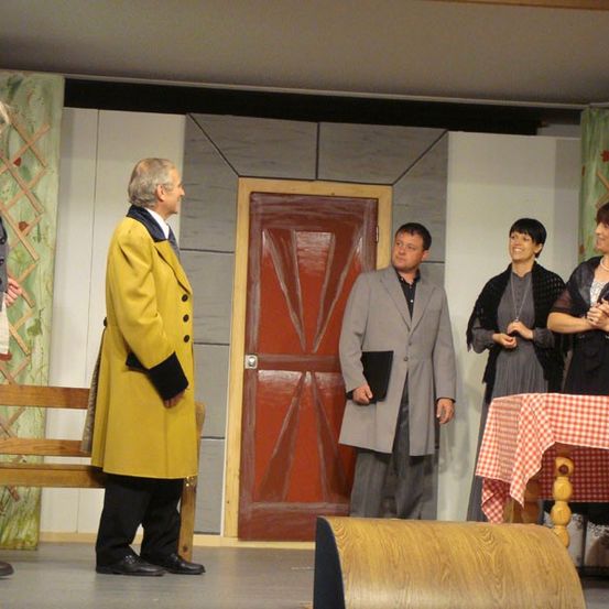 Fünf Personen stehen auf der Bühne mit einer roten Tür und einer Bank hinter ihnen. Der Mann im gelben Mantel hält ein Buch.