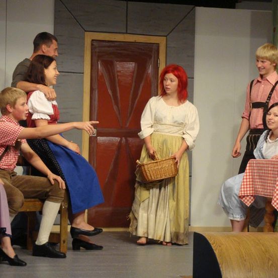 Eine Gruppe von Schauspielern führt ein Theaterstück auf der Bühne auf, wobei eine Frau einen Korb hält und eine andere Frau auf sie zeigt.