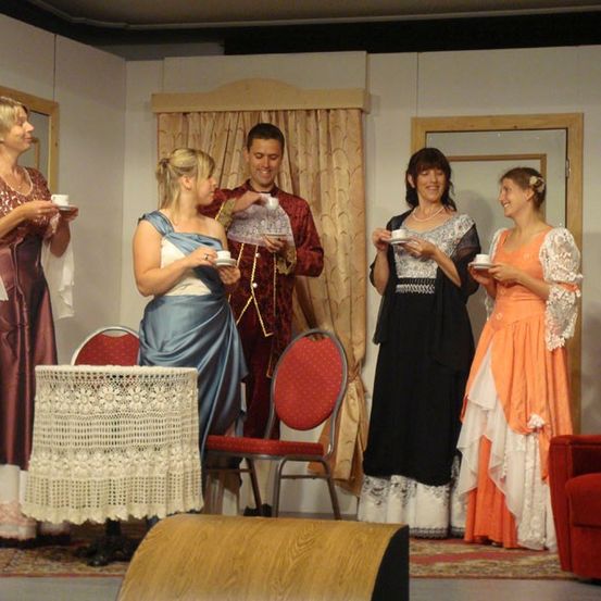 Fünf Personen sind drinnen, in Kostümen gekleidet, möglicherweise für ein Theaterstück probend. Zwei Frauen halten Tassen und Untertassen, während ein Mann zwischen ihnen steht.