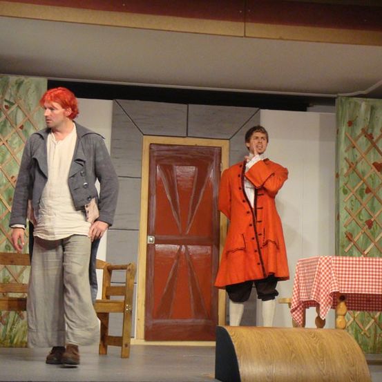 Zwei Männer stehen auf der Bühne, einer in einem grauen Mantel und der andere in einem roten Mantel. Hinter ihnen steht eine Holztür mit einem Tisch in der Nähe.