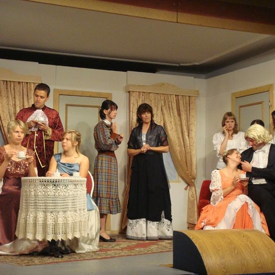 Eine Gruppe von Schauspielern in historischen Kostümen steht auf der Bühne. Einige sitzen um einen Tisch herum, während andere dahinter stehen. Zwei Personen unterhalten sich rechts.